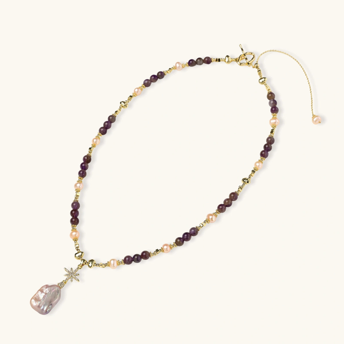 Gold necklace with iridescent baroque pearl pendant, purple amethyst beads, and crystal star charm on cream background